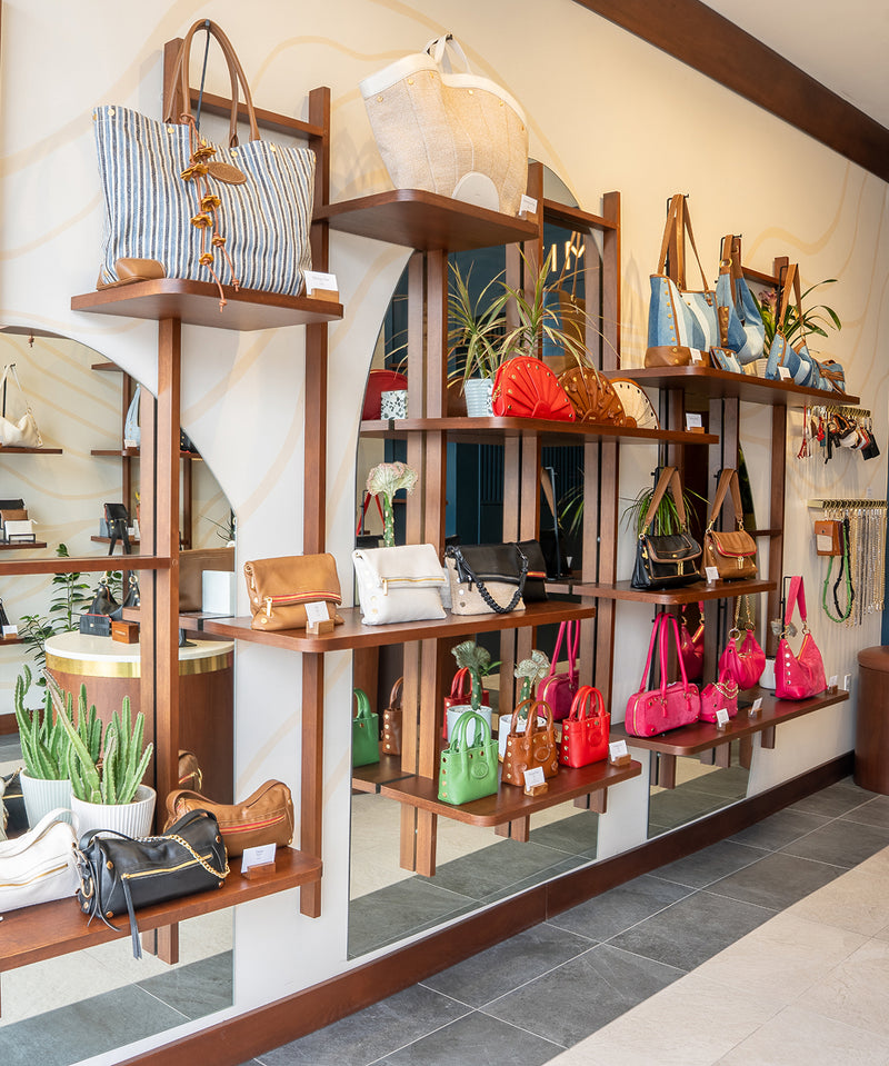 A retail display of multiple handbags and tote bags arranged on wooden shelving units inside a boutique. The shelves feature a variety of colors and patterns, including blue and white stripes, solid red, green, pink, and brown, with decorative plants and mirrors.
