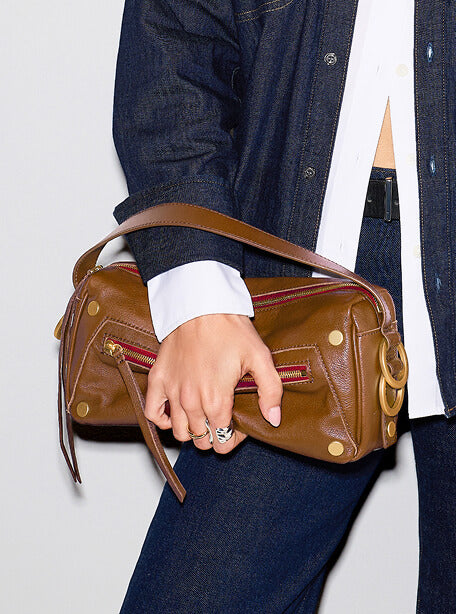 A person wearing a dark denim jacket and jeans holds a brown leather shoulder bag with gold-tone hardware and red zipper accents.