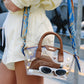 A clear acrylic handbag with brown leather handles and trim, styled with a blue patterned scarf strap, gold studs, and accessories inside, carried by a person wearing a cream outfit.