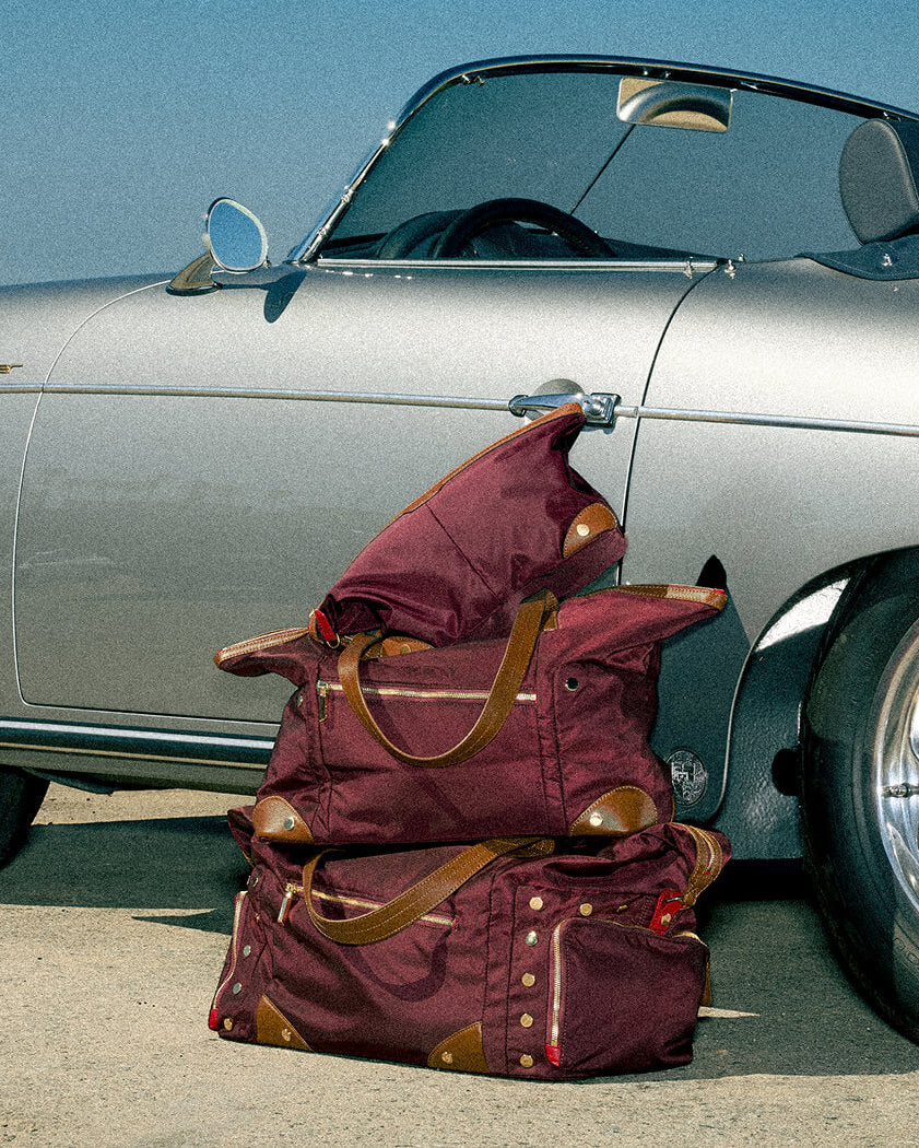 A stack of burgundy and brown canvas duffel bags sits on the ground next to a silver convertible car under a clear blue sky.