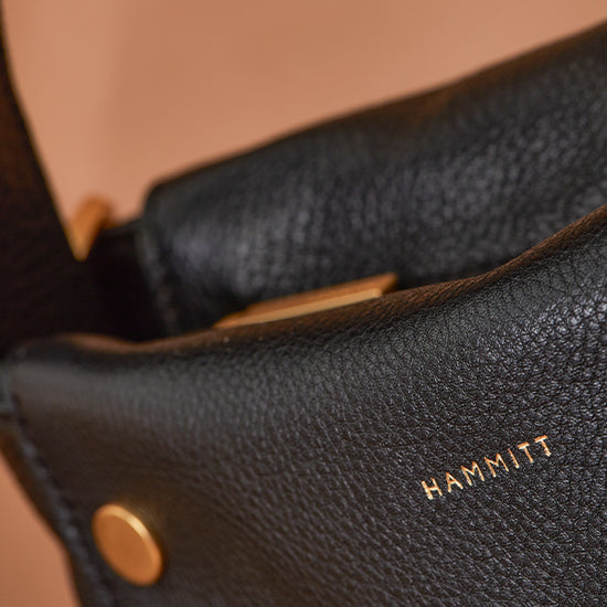 Image: Close up up Hammitt logo in gold on black handbag.