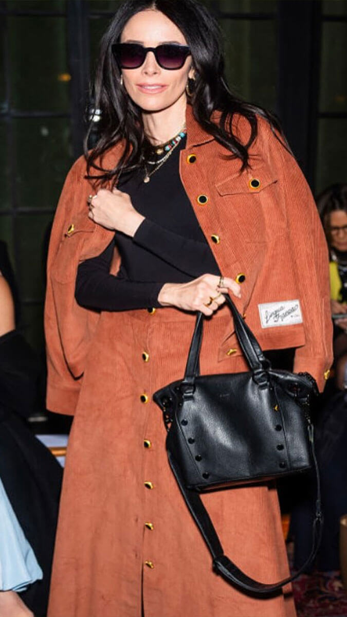 A woman wearing a rust-orange corduroy coat with gold buttons over a black top, accessorized with a black studded handbag and sunglasses.