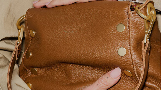 A close-up of a brown pebbled leather shoulder bag with gold-tone hardware, held by hands against a beige fabric background.