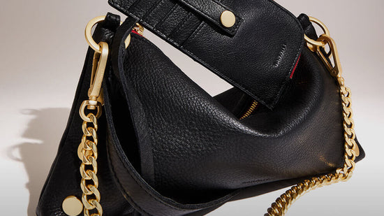 A close-up of a black pebbled leather handbag with gold-tone hardware, including a chain strap, gold snap button, and a small red accent detail visible inside the bag.