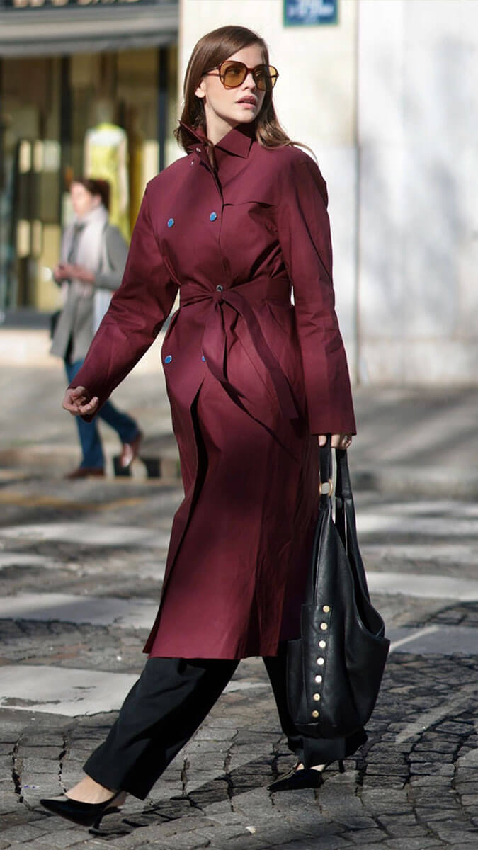 A woman walking outdoors on a cobblestone street wearing a long burgundy trench coat with a tie waist, black pants, black pointed shoes, and carrying a large black shoulder bag.