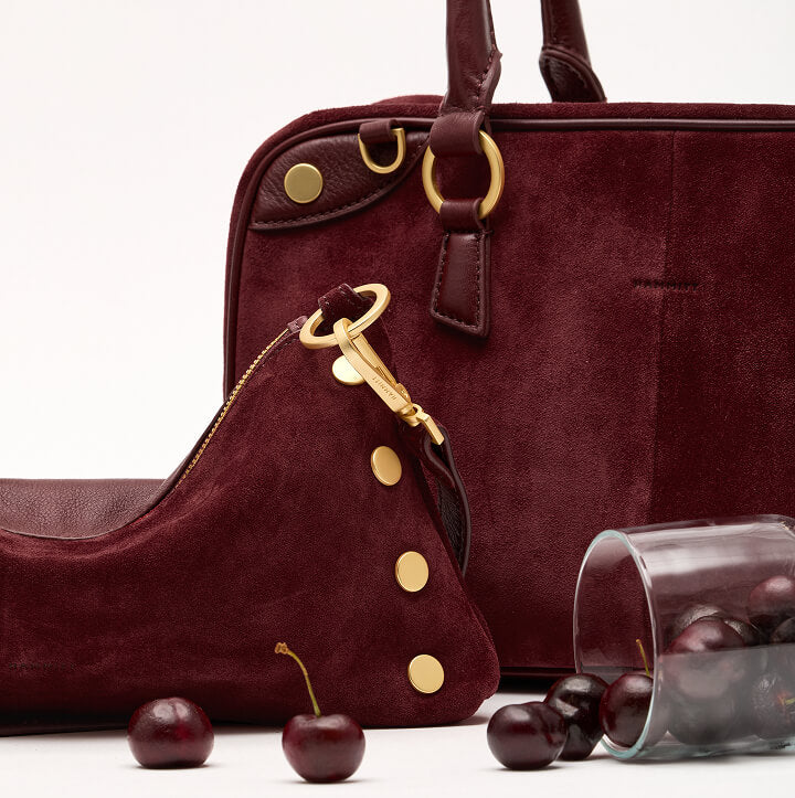 A burgundy suede and leather handbag set with gold-tone hardware, shown on a clean white background with a few cherries and a glass of cherries as styling props.