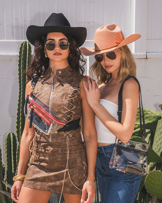 Image of two models in festival attire in front of white wall and cacti. One model has charles clear gold red zip and the other has Tony Sml clear in black gunmetal.