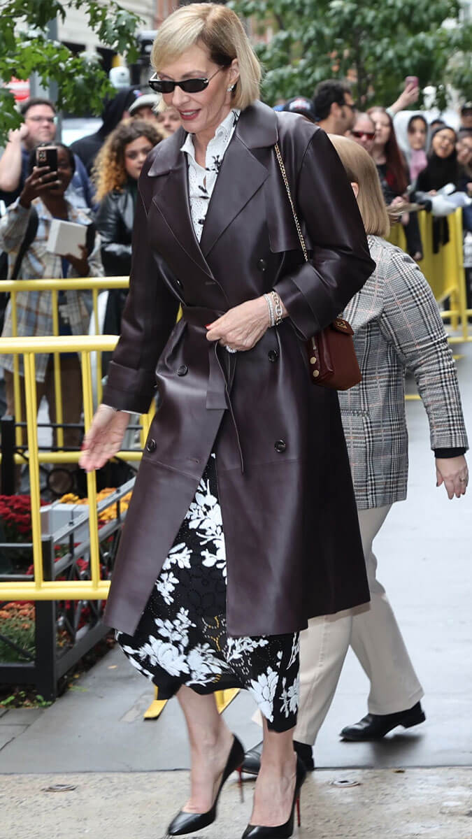 A woman walking outdoors wearing a long dark brown leather trench coat over a black and white floral dress, with black heels and a brown shoulder bag.