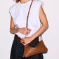 A woman wearing a white sleeveless top and dark jeans carries a brown leather shoulder bag with gold-tone hardware and a thin brown strap.