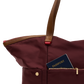 A close-up of a burgundy nylon tote bag with brown leather handles and a front zip pocket holding a navy blue passport, set against a plain white background.