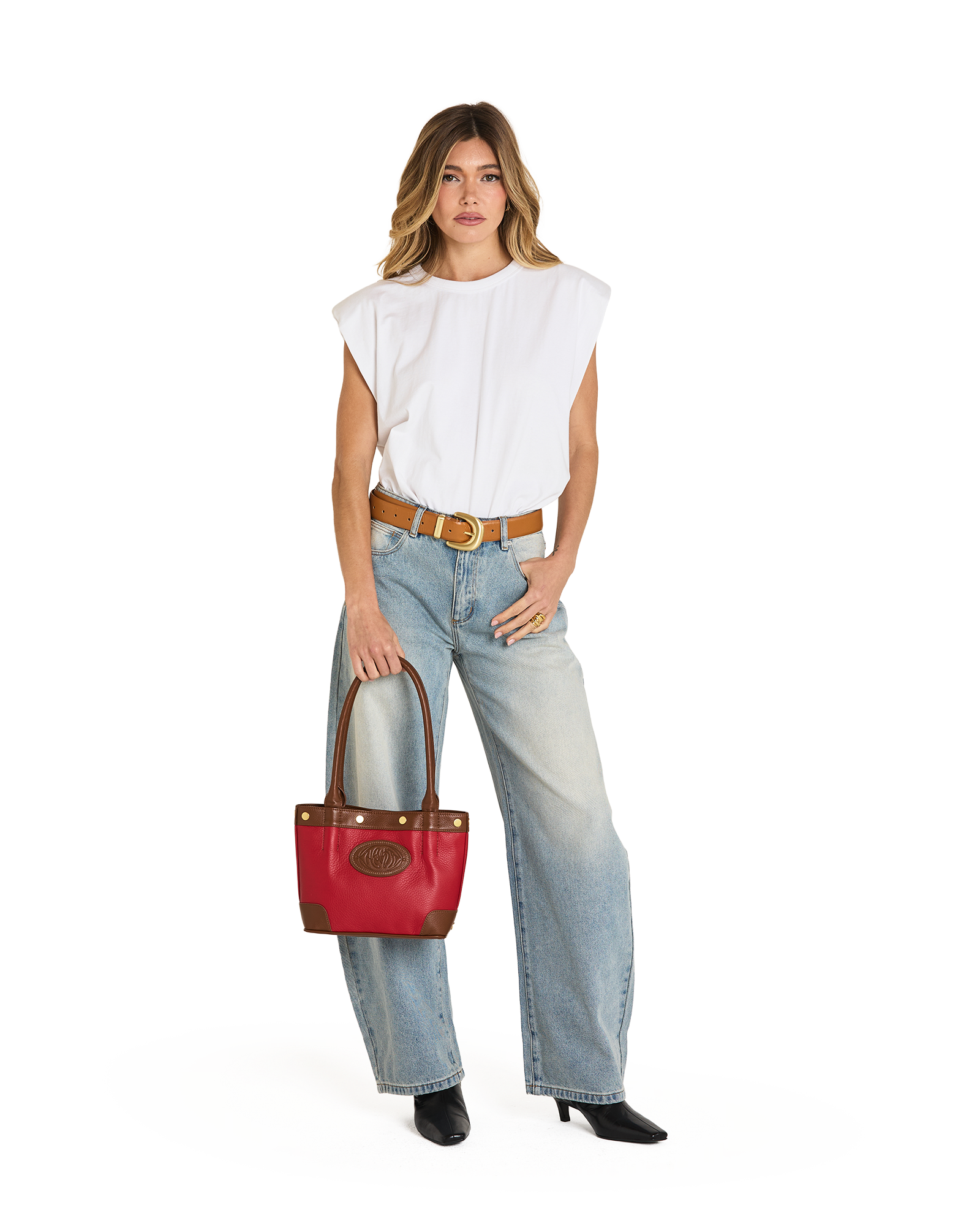 A woman modeling a white sleeveless top, light-wash wide-leg jeans, a brown belt, and black boots, holding a red leather handbag with brown leather handles and trim against a plain white background.