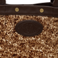 Close-up of a handbag featuring a brown and beige cowhide patterned body, dark brown leather trim with gold-tone rivets, and a dark brown oval embossed logo patch.