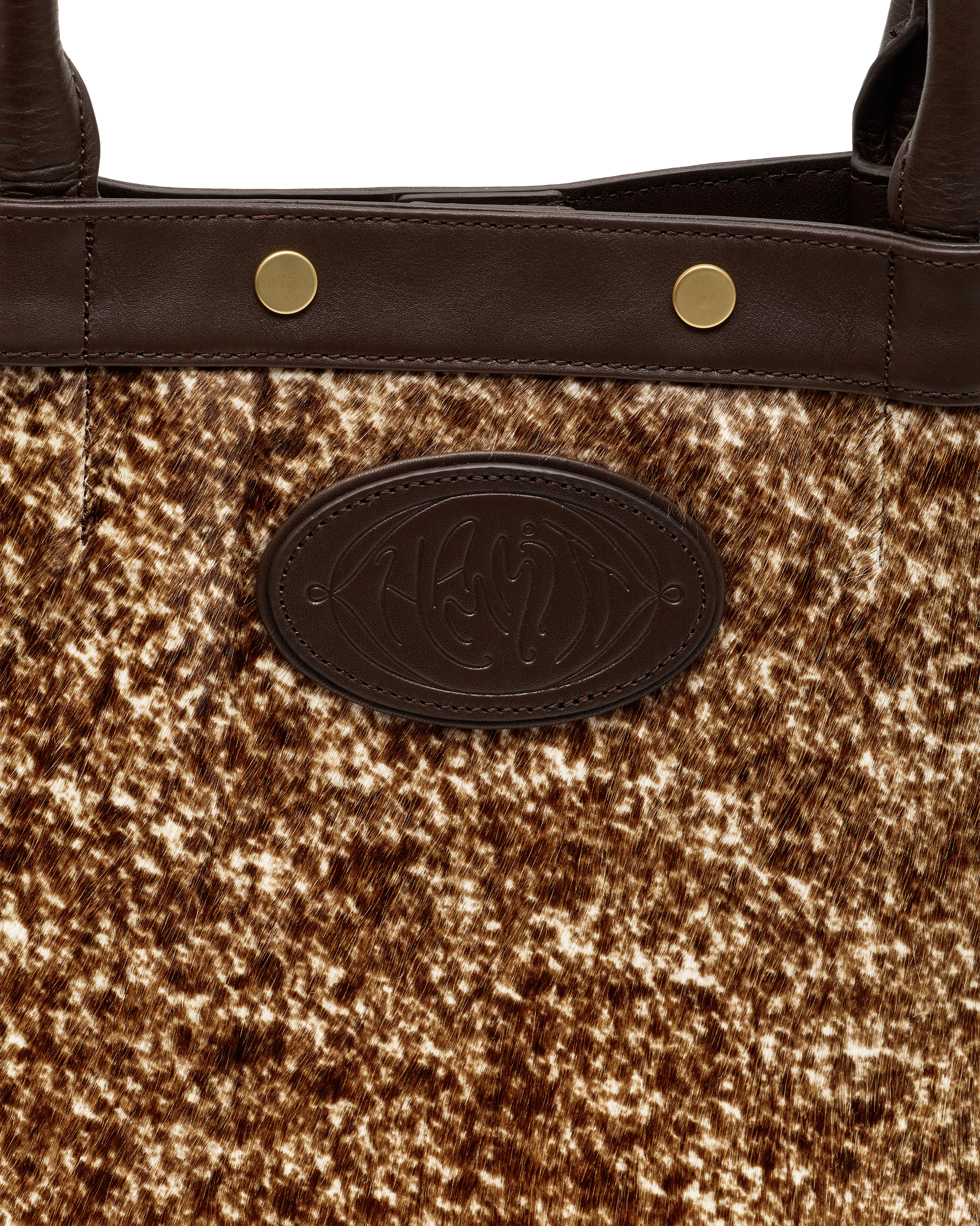 Close-up of a handbag featuring a brown and beige cowhide patterned body, dark brown leather trim with gold-tone rivets, and a dark brown oval embossed logo patch.