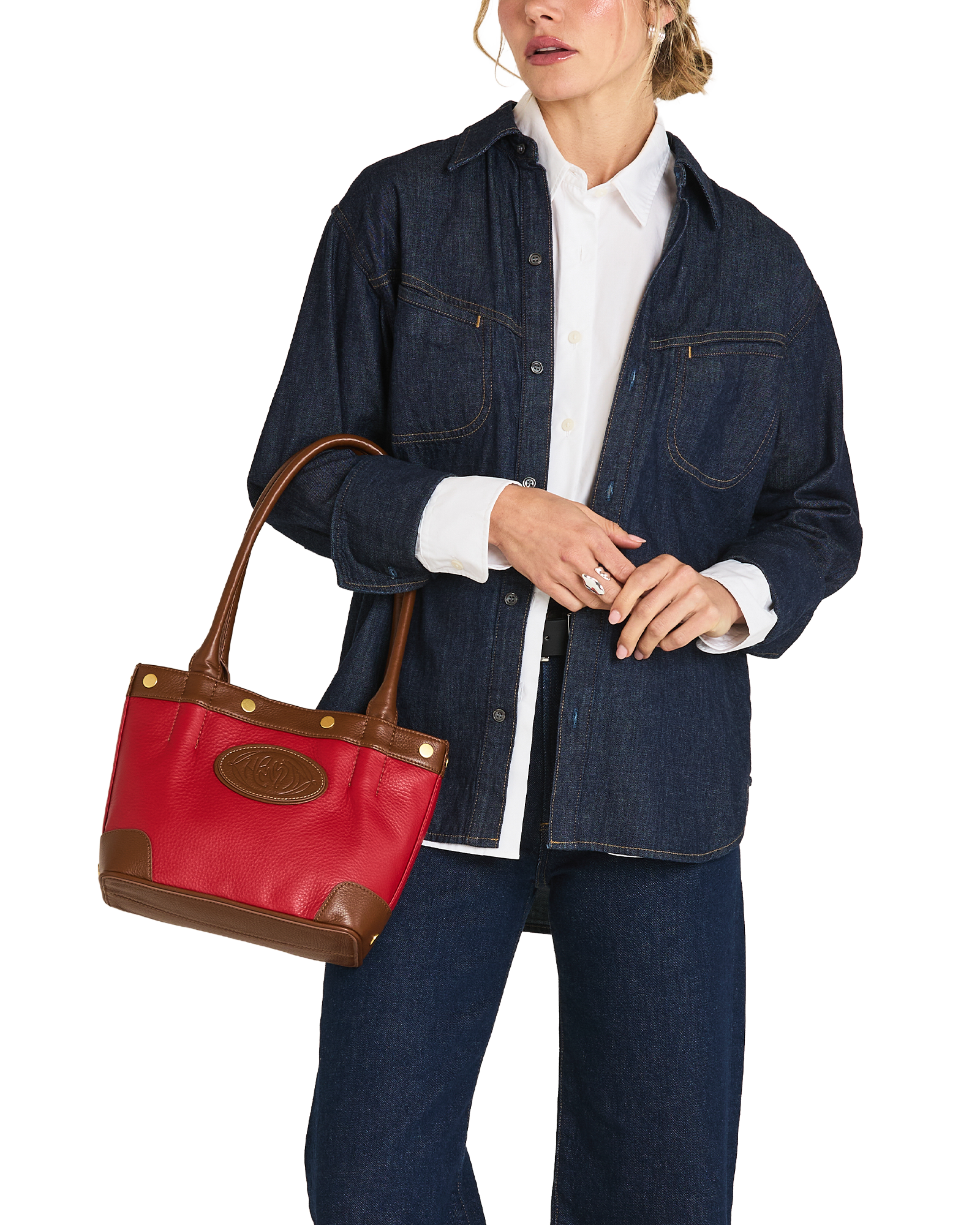 A model wearing a dark blue denim shirt jacket, white button-down shirt, and dark blue jeans, holding a red leather tote bag with brown leather handles and trim.