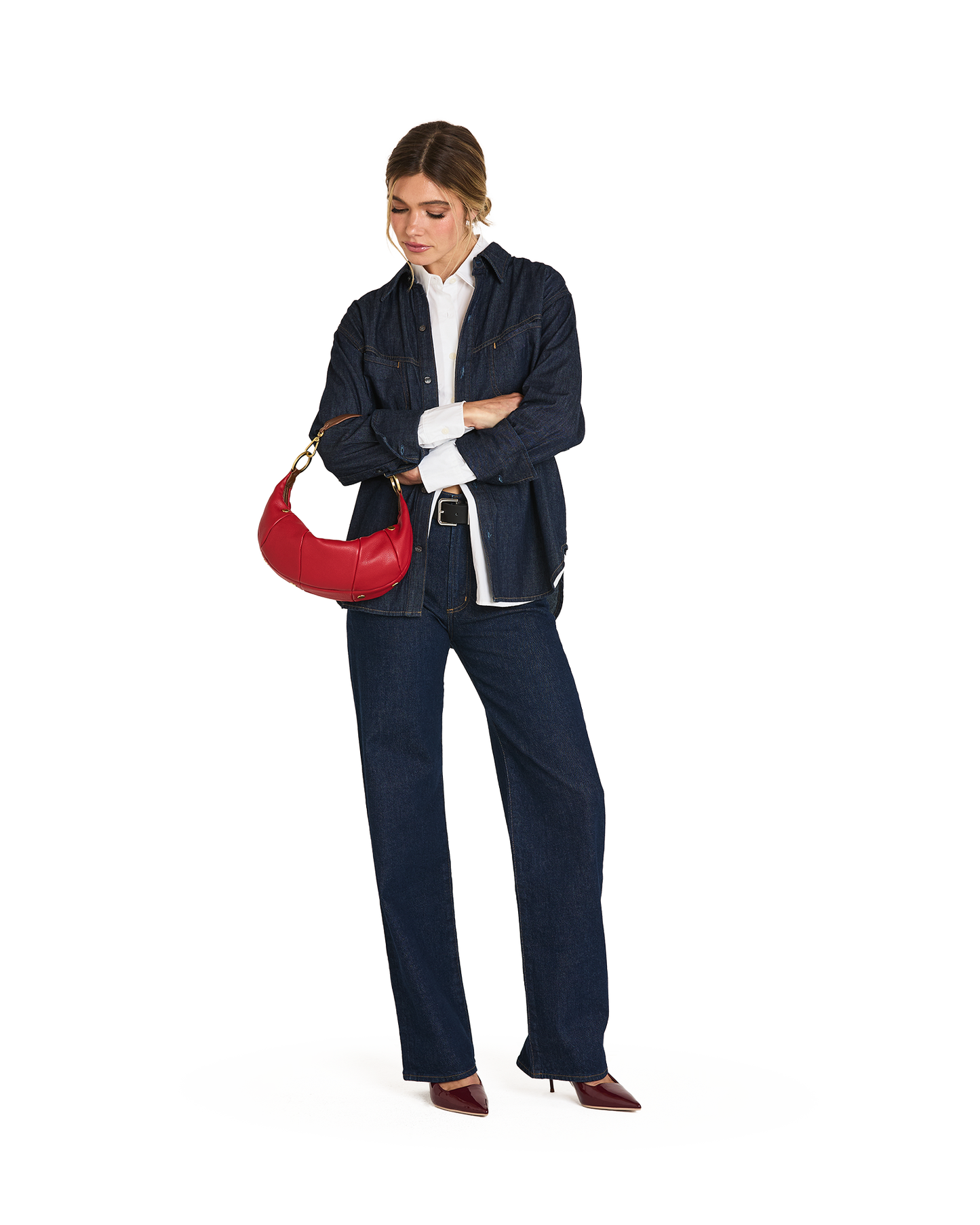 A woman modeling a dark navy blue denim jacket and matching wide-leg jeans, styled with a white collared shirt, dark red pointed-toe heels, and a red crescent shoulder bag against a plain white background.