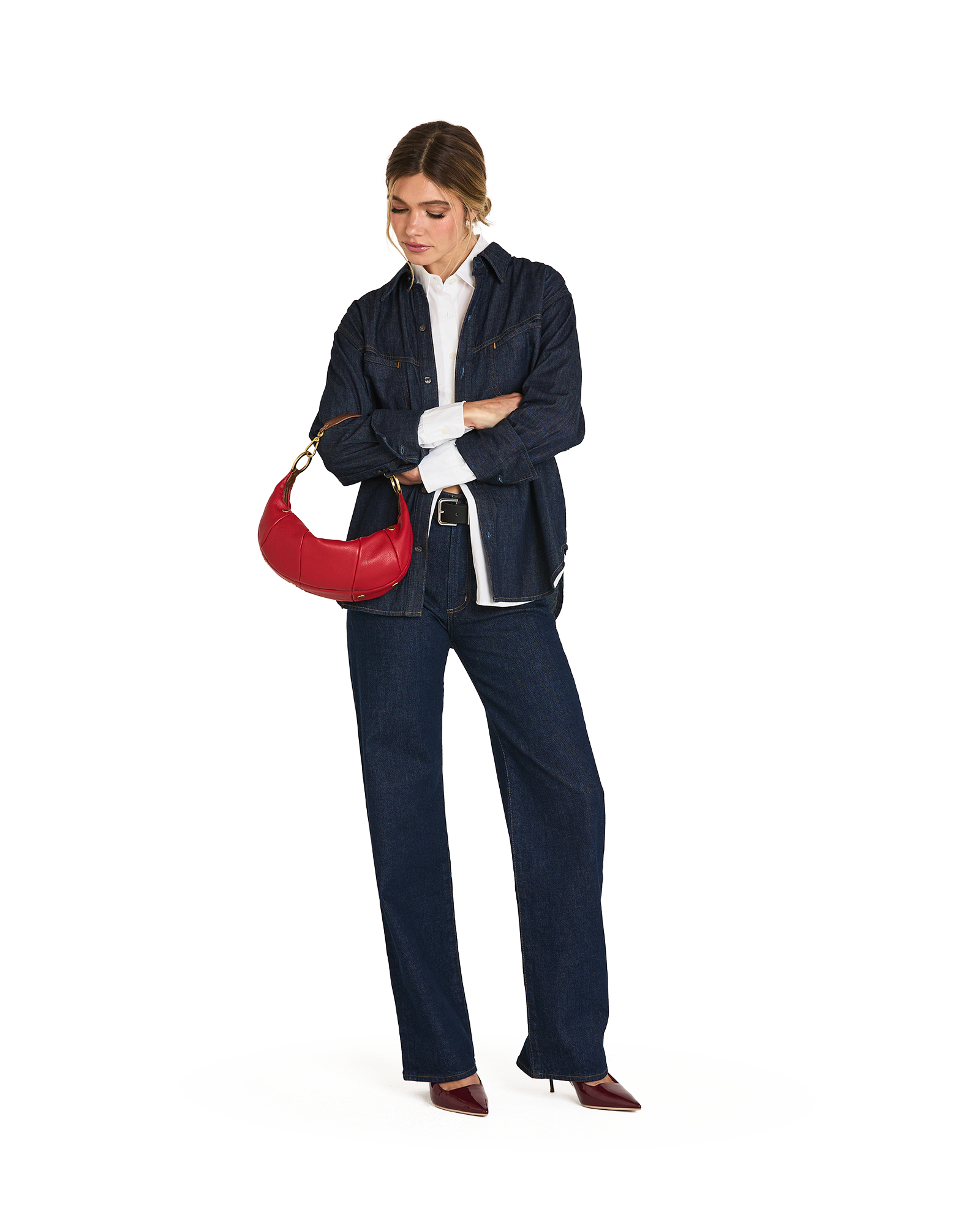 A woman modeling a dark navy blue denim jacket and matching wide-leg jeans, styled with a white collared shirt, dark red pointed-toe heels, and a red crescent shoulder bag against a plain white background.