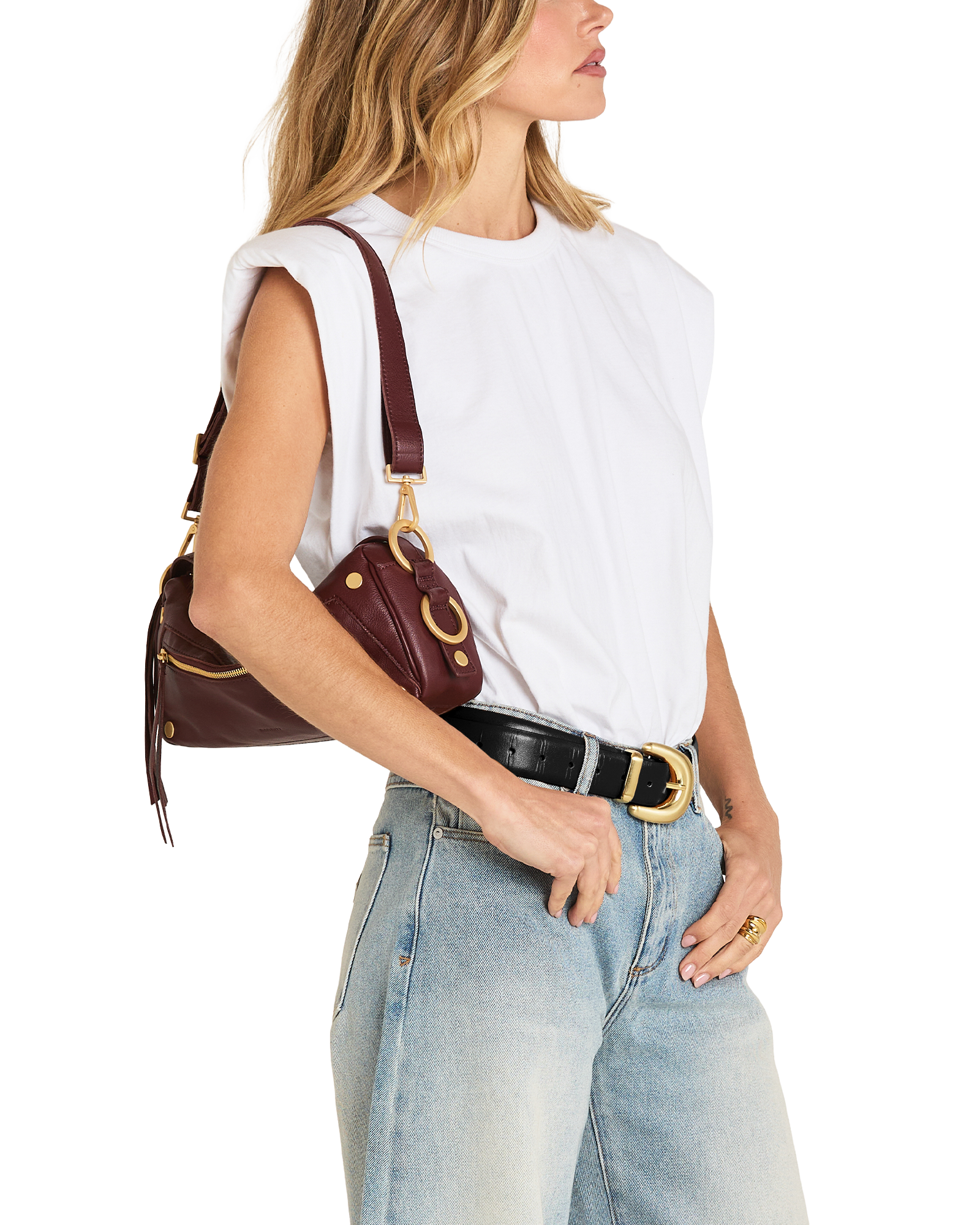 A woman wearing a white sleeveless top, light blue jeans, and a black belt with a gold buckle, carrying a dark burgundy leather shoulder bag with gold hardware.