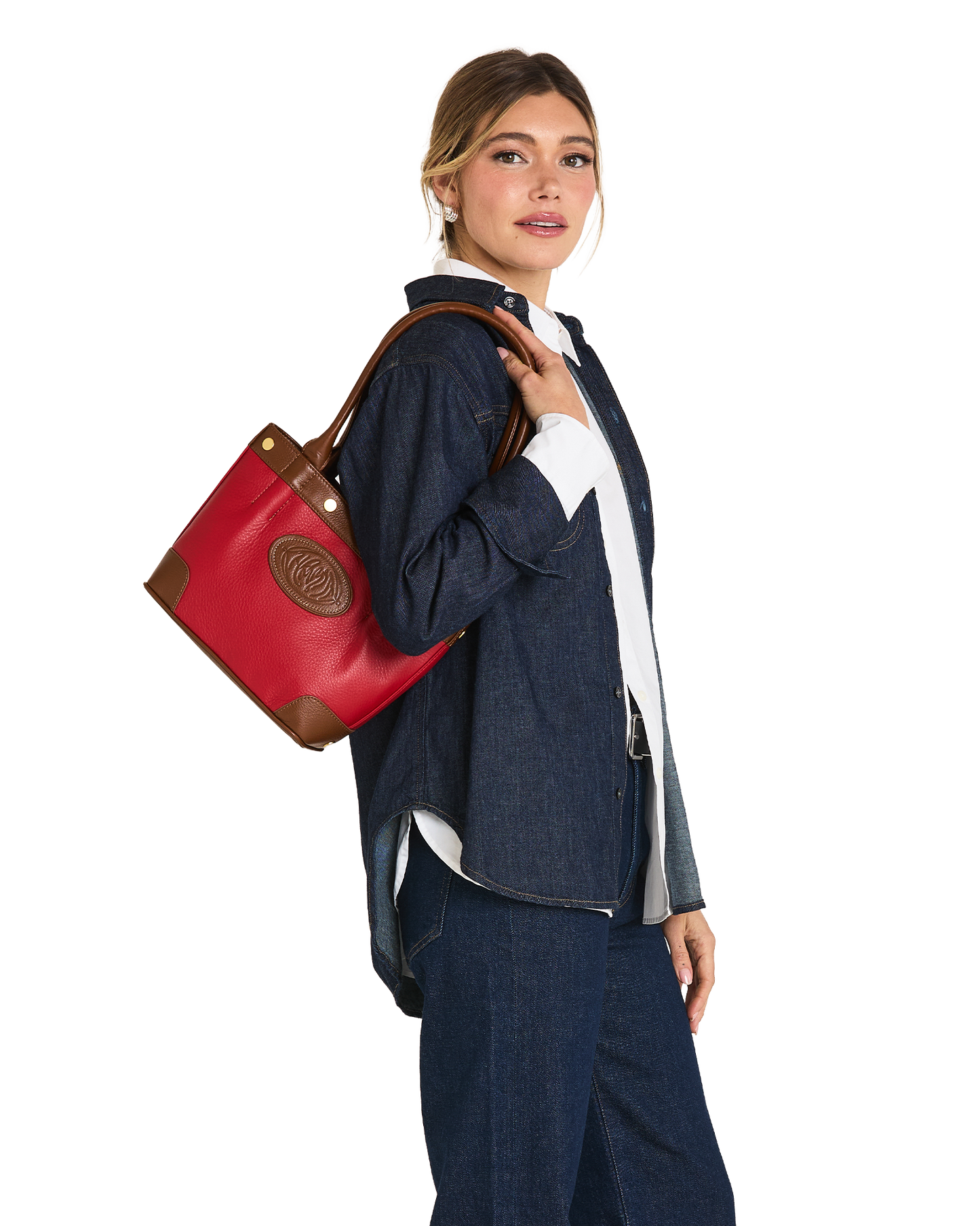 A woman in a denim outfit carries a red leather tote bag with brown trim and a circular logo patch on a plain white background.
