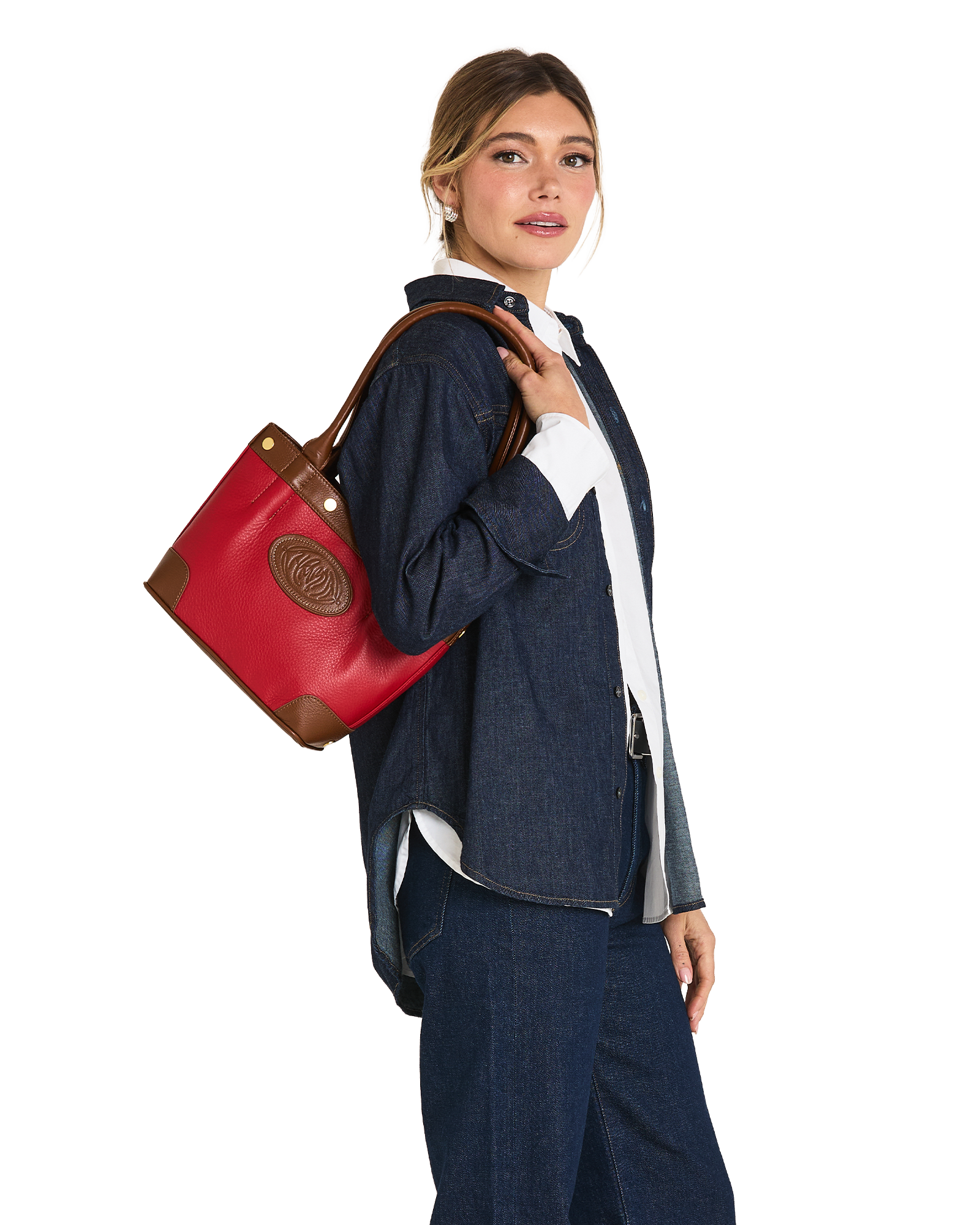 A woman in a denim outfit carries a red leather tote bag with brown trim and a circular logo patch on a plain white background.
