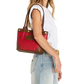 A woman in a white sleeveless top and light blue jeans carries a red and brown leather tote bag over her shoulder, shown against a plain white background.