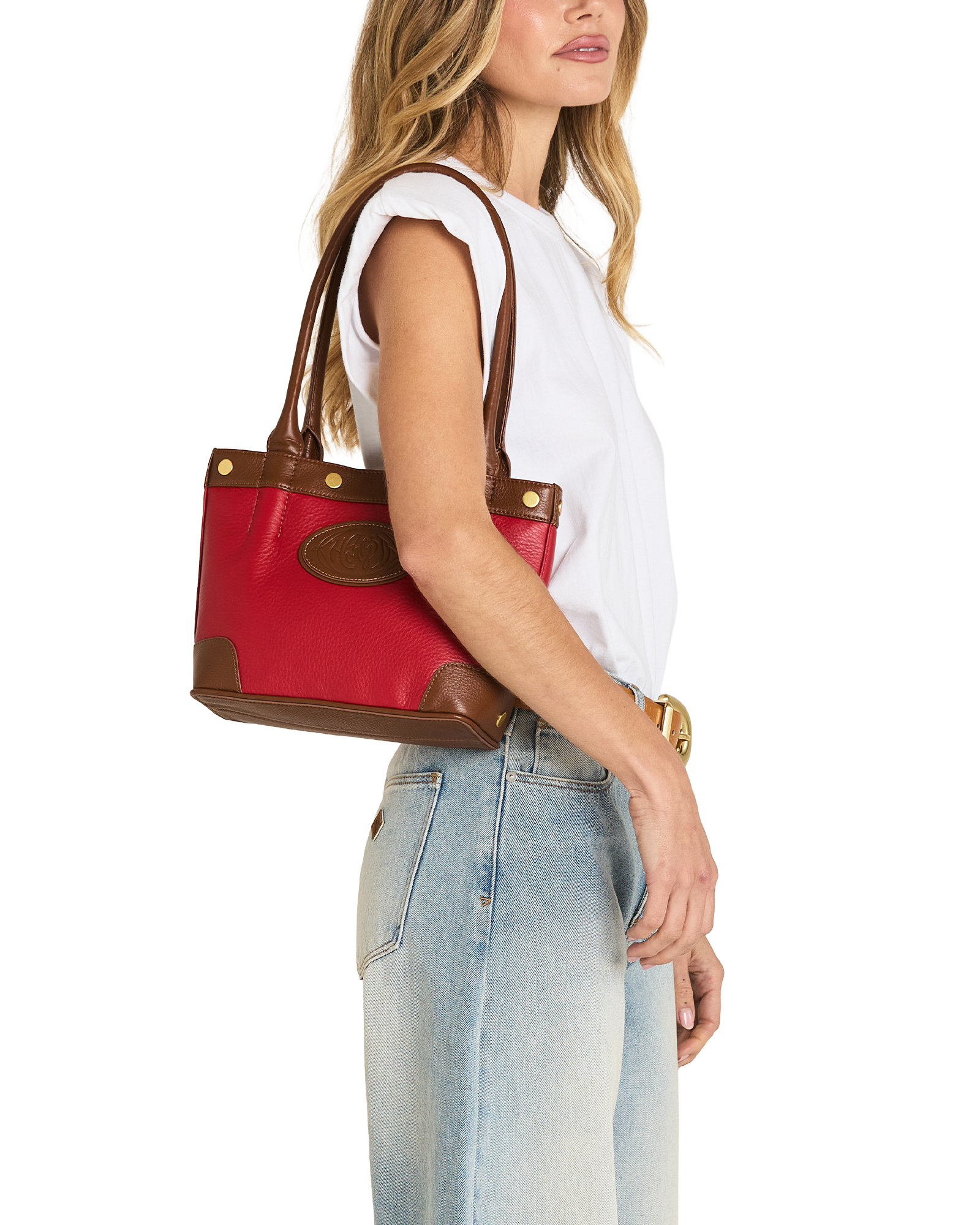 A woman in a white sleeveless top and light blue jeans carries a red and brown leather tote bag over her shoulder, shown against a plain white background.
