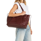 A woman wearing a white sleeveless top and light blue jeans carries a large burgundy leather tote bag with gold-tone studs and a shoulder strap against a plain white background.