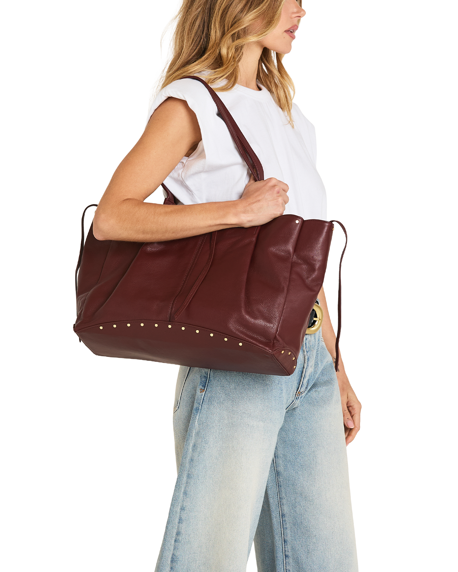 A woman wearing a white sleeveless top and light blue jeans carries a large burgundy leather tote bag with gold-tone studs and a shoulder strap against a plain white background.