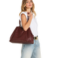 A woman wearing a white sleeveless top, light blue jeans, and a gold-buckled belt carries a large burgundy leather tote bag with gold-tone studs on a plain white background.