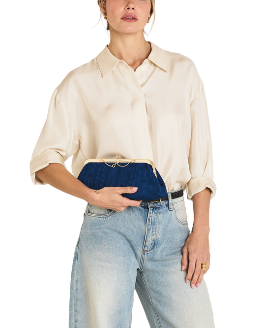 A model wearing a cream-colored silk shirt and light-wash blue jeans holds a navy blue velvet clutch with a gold-tone kiss-lock closure.