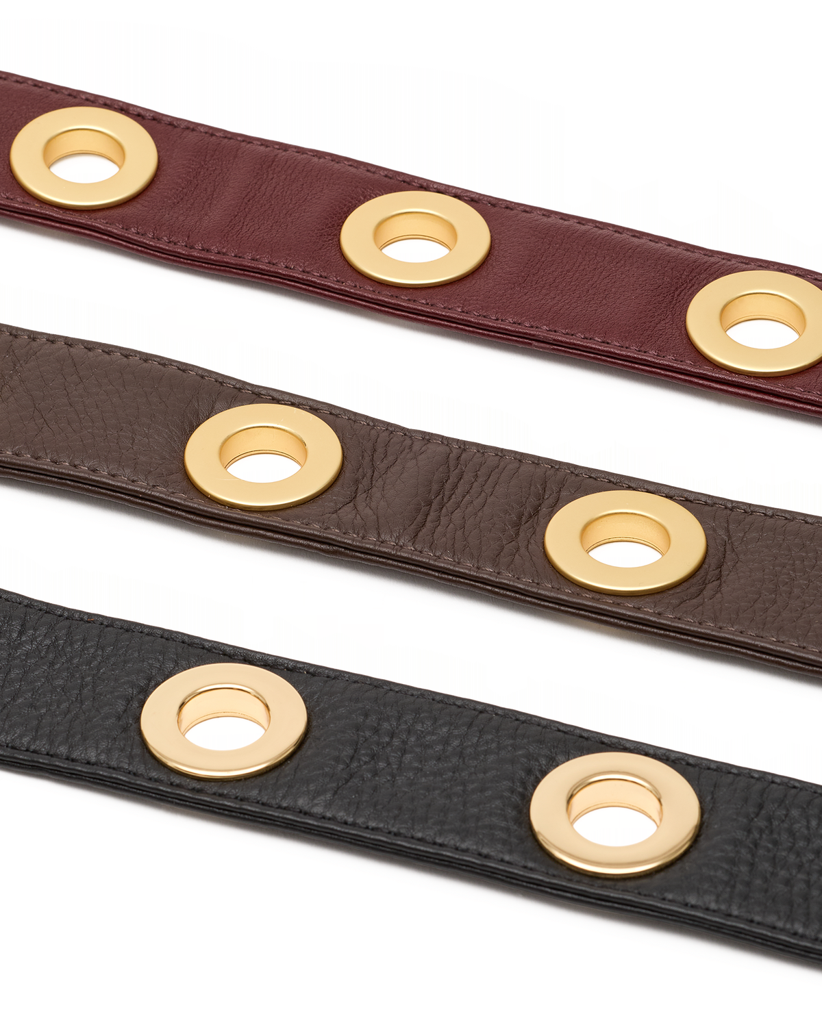 Three leather straps in burgundy, brown, and black, each featuring a row of polished gold-tone circular grommets, displayed diagonally on a white background.