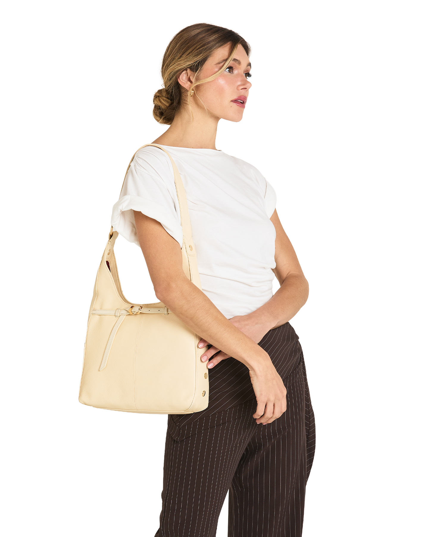 A woman wearing a white t-shirt and dark brown pinstriped trousers carries a light beige leather shoulder bag on her shoulder, shown from a side profile against a solid white background.