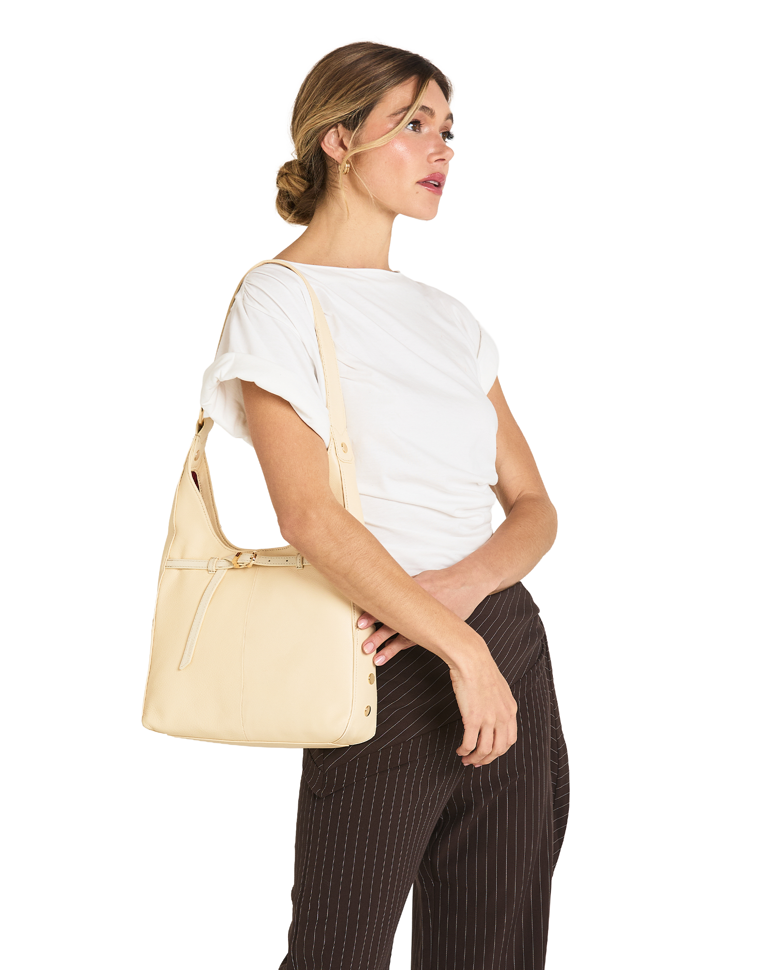 A woman wearing a white t-shirt and dark brown pinstriped trousers carries a light beige leather shoulder bag on her shoulder, shown from a side profile against a solid white background.