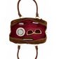 An open brown leather handbag with a deep red interior, shown against a plain white background. The bag contains several accessories, including beige-framed sunglasses, a white round cosmetic case, and a small dark rectangular item.