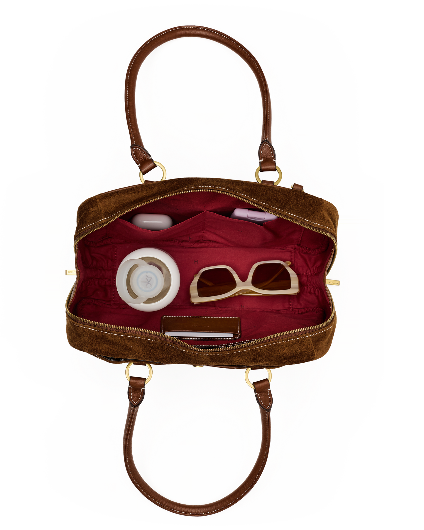 An open brown leather handbag with a deep red interior, shown against a plain white background. The bag contains several accessories, including beige-framed sunglasses, a white round cosmetic case, and a small dark rectangular item.