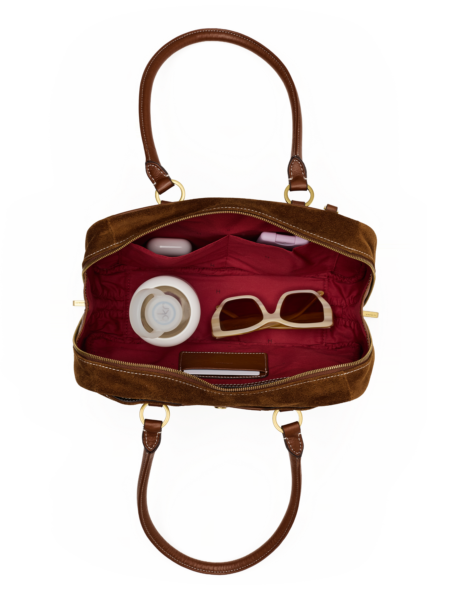 An open brown leather handbag with a deep red interior, shown against a plain white background. The bag contains several accessories, including beige-framed sunglasses, a white round cosmetic case, and a small dark rectangular item.