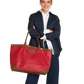 A woman stands against a white background holding a large red leather tote bag with brown trim and handles. She is wearing a dark denim jacket over a white collared shirt and dark jeans.