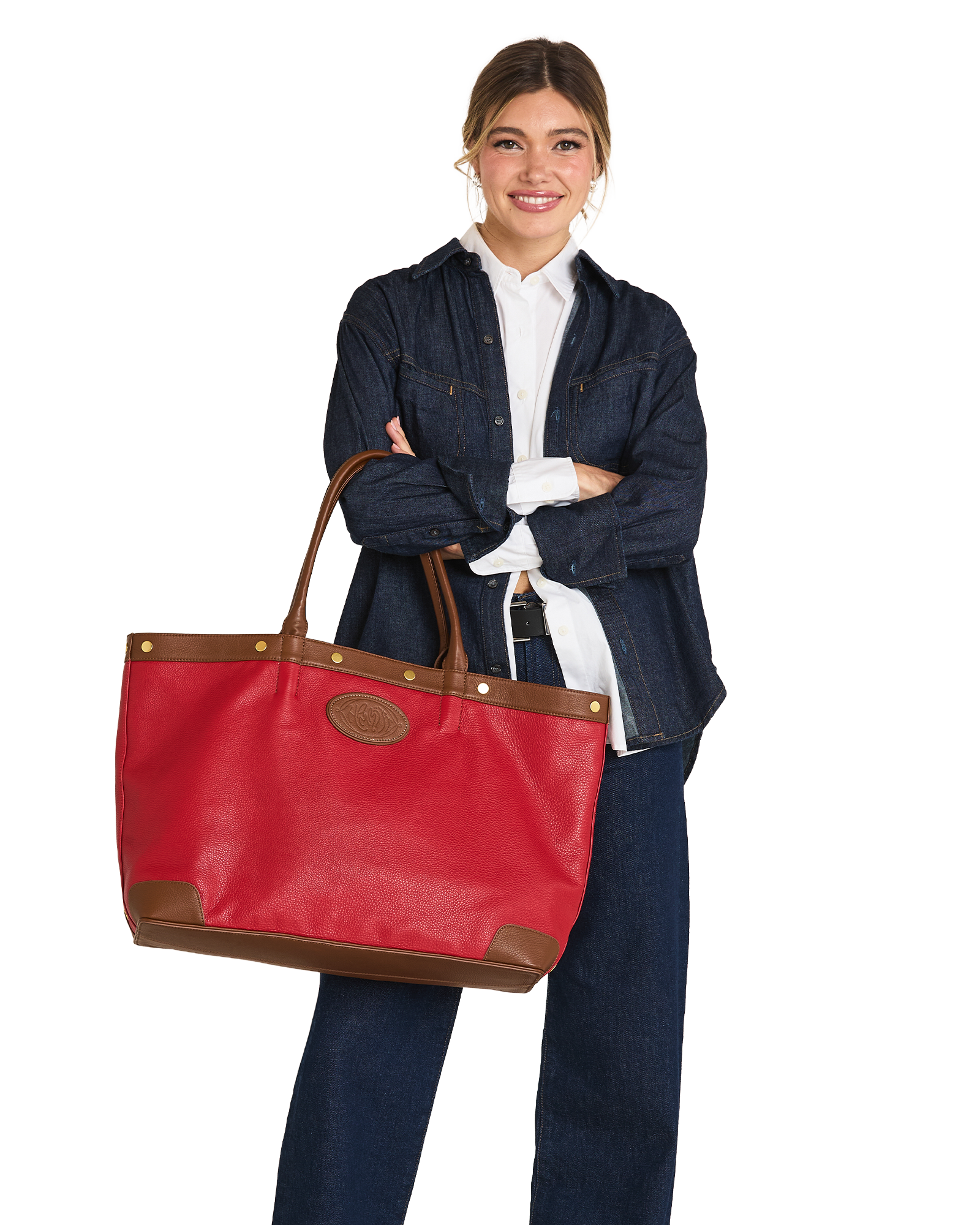 A woman stands against a white background holding a large red leather tote bag with brown trim and handles. She is wearing a dark denim jacket over a white collared shirt and dark jeans.