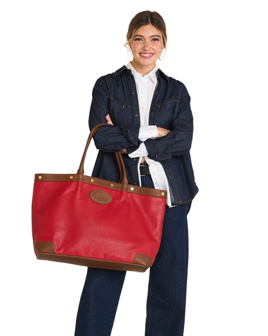A woman stands against a white background holding a large red leather tote bag with brown trim and handles. She is wearing a dark denim jacket over a white collared shirt and dark jeans.