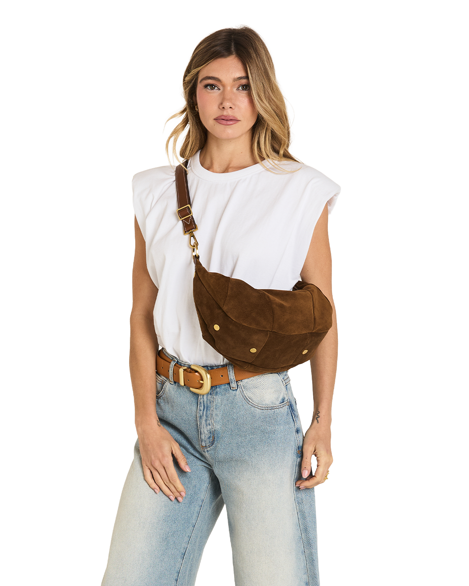 A woman wearing a brown suede crescent-shaped crossbody bag with gold-tone hardware, styled with a white sleeveless top and light blue jeans against a white background.
