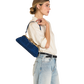 A woman in a cream shirt and light blue jeans carries a blue velvet shoulder bag with gold hardware against a plain white background.