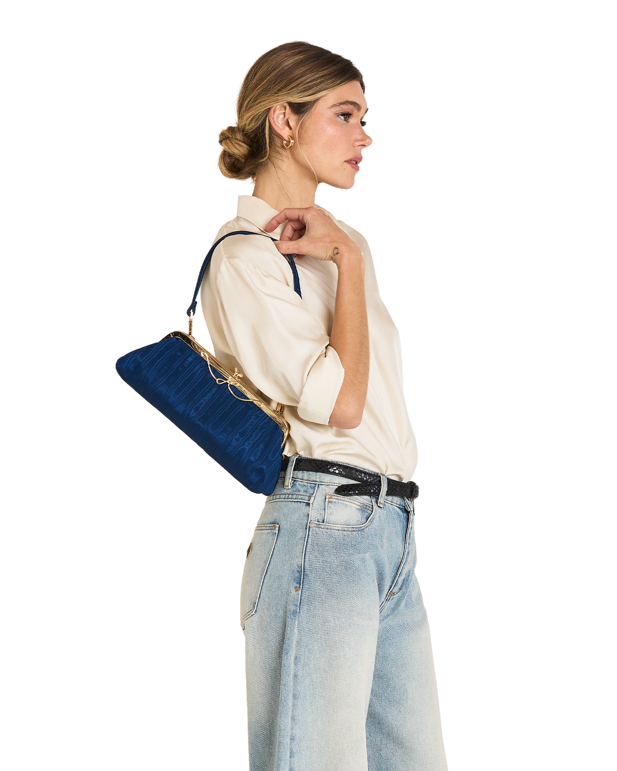 A woman in a cream shirt and light blue jeans carries a blue velvet shoulder bag with gold hardware against a plain white background.