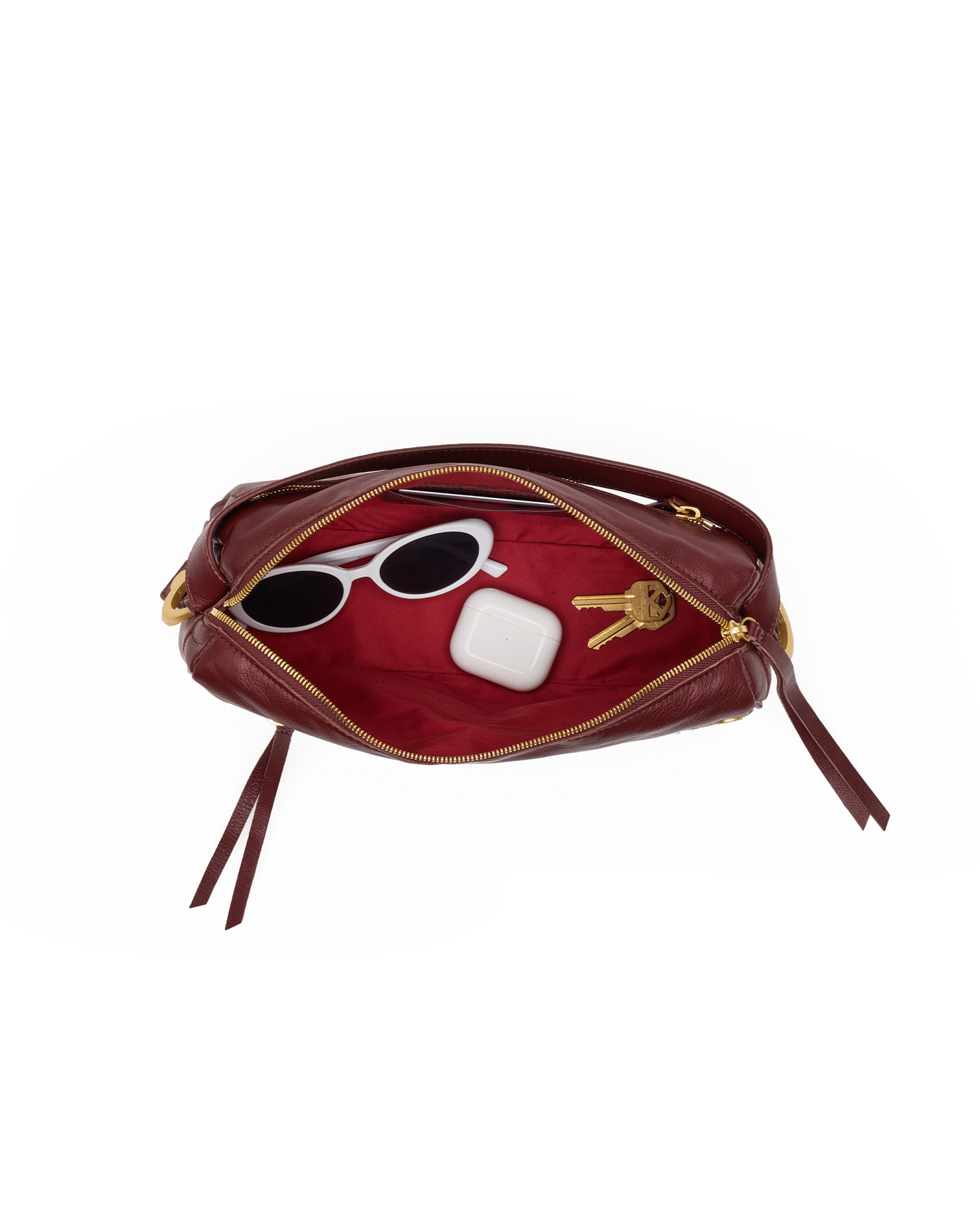 A burgundy leather pouch shown open on a white background, revealing a red interior with gold-tone zipper hardware. White sunglasses, a white earbud case, and two keys are placed inside as styling props.