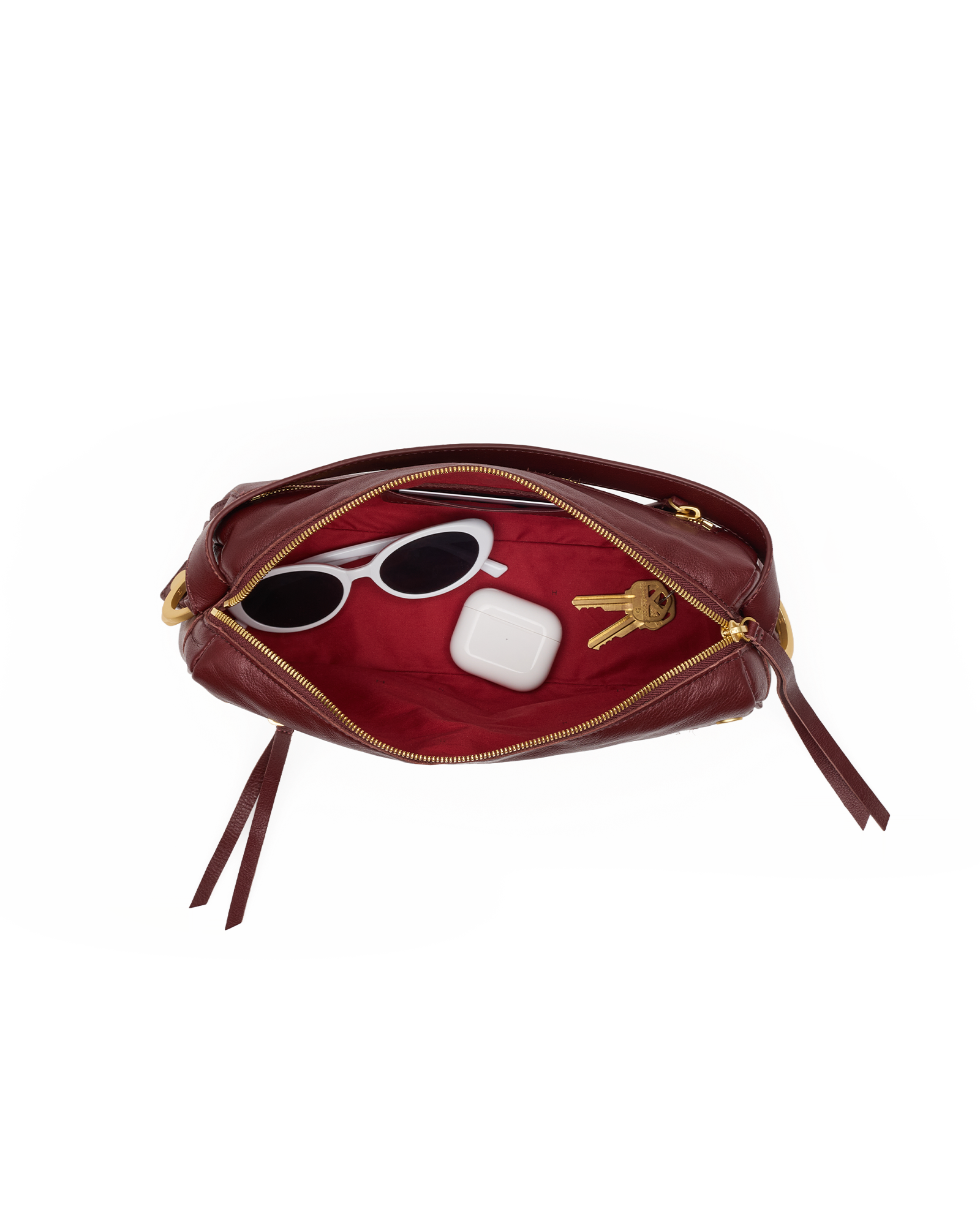 A burgundy leather pouch shown open on a white background, revealing a red interior with gold-tone zipper hardware. White sunglasses, a white earbud case, and two keys are placed inside as styling props.
