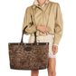 A model wearing a beige trench coat and cream shorts holds a large brown glittery tote bag with dark brown leather trim and handles against a plain white background.