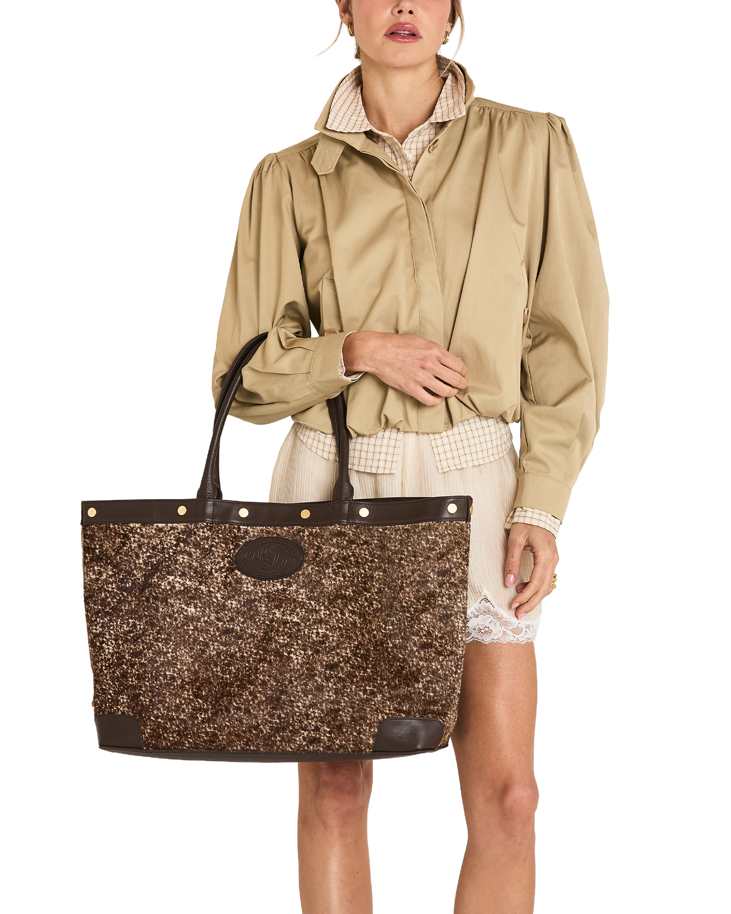 A model wearing a beige trench coat and cream shorts holds a large brown glittery tote bag with dark brown leather trim and handles against a plain white background.