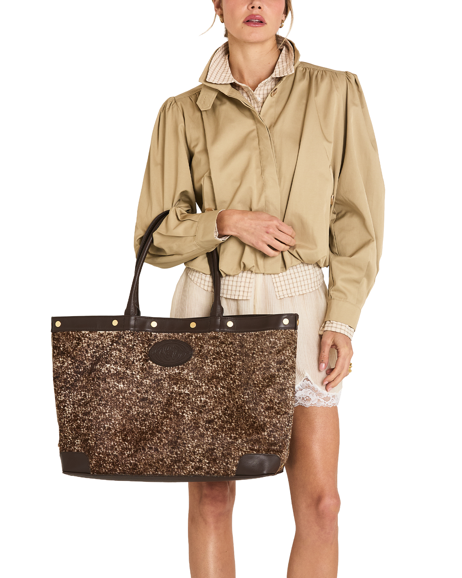 A model wearing a beige trench coat and cream shorts holds a large brown glittery tote bag with dark brown leather trim and handles against a plain white background.
