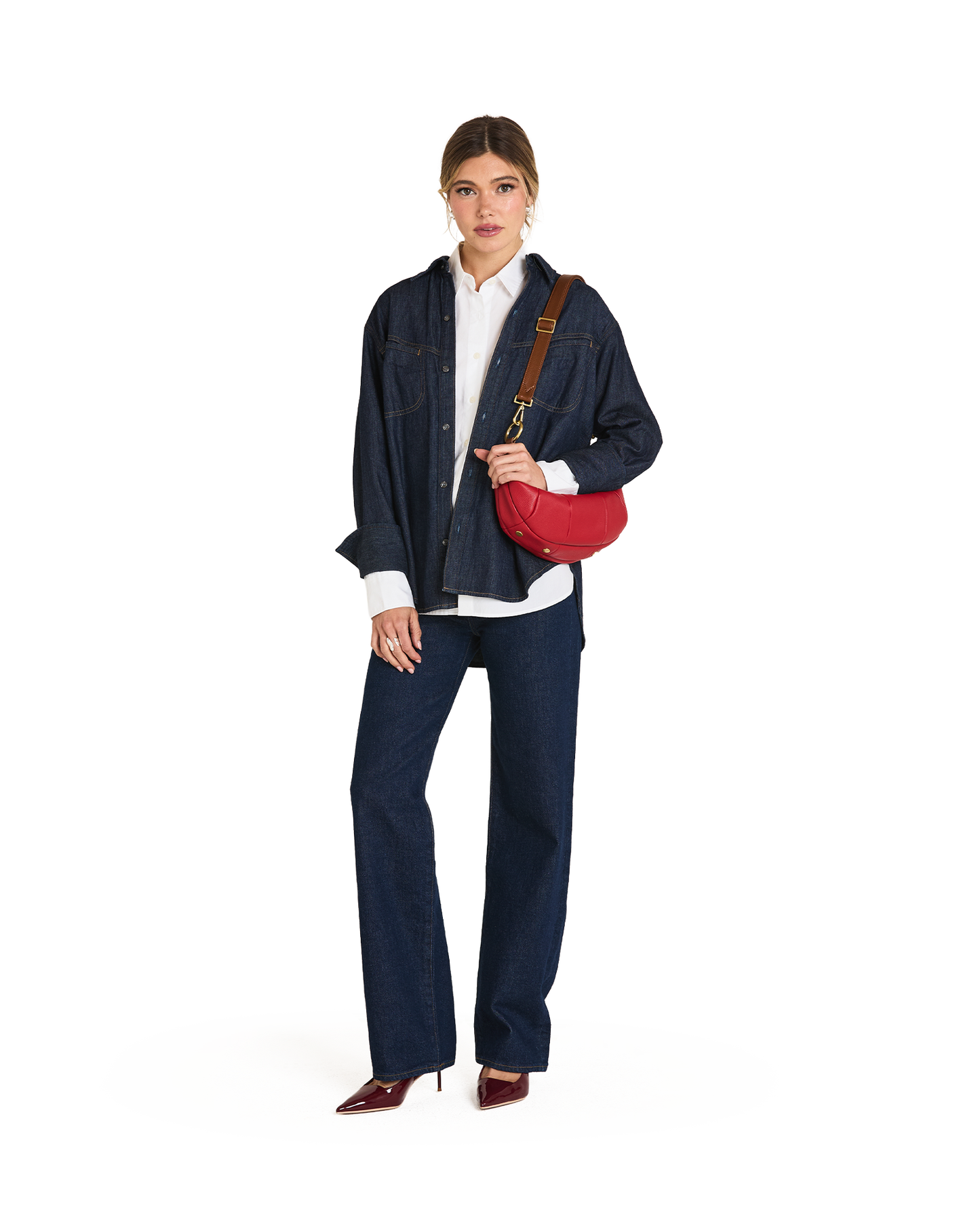 A model stands against a white studio background wearing a dark blue denim jacket and matching wide-leg jeans over a white shirt, with a red crossbody bag and brown heels.