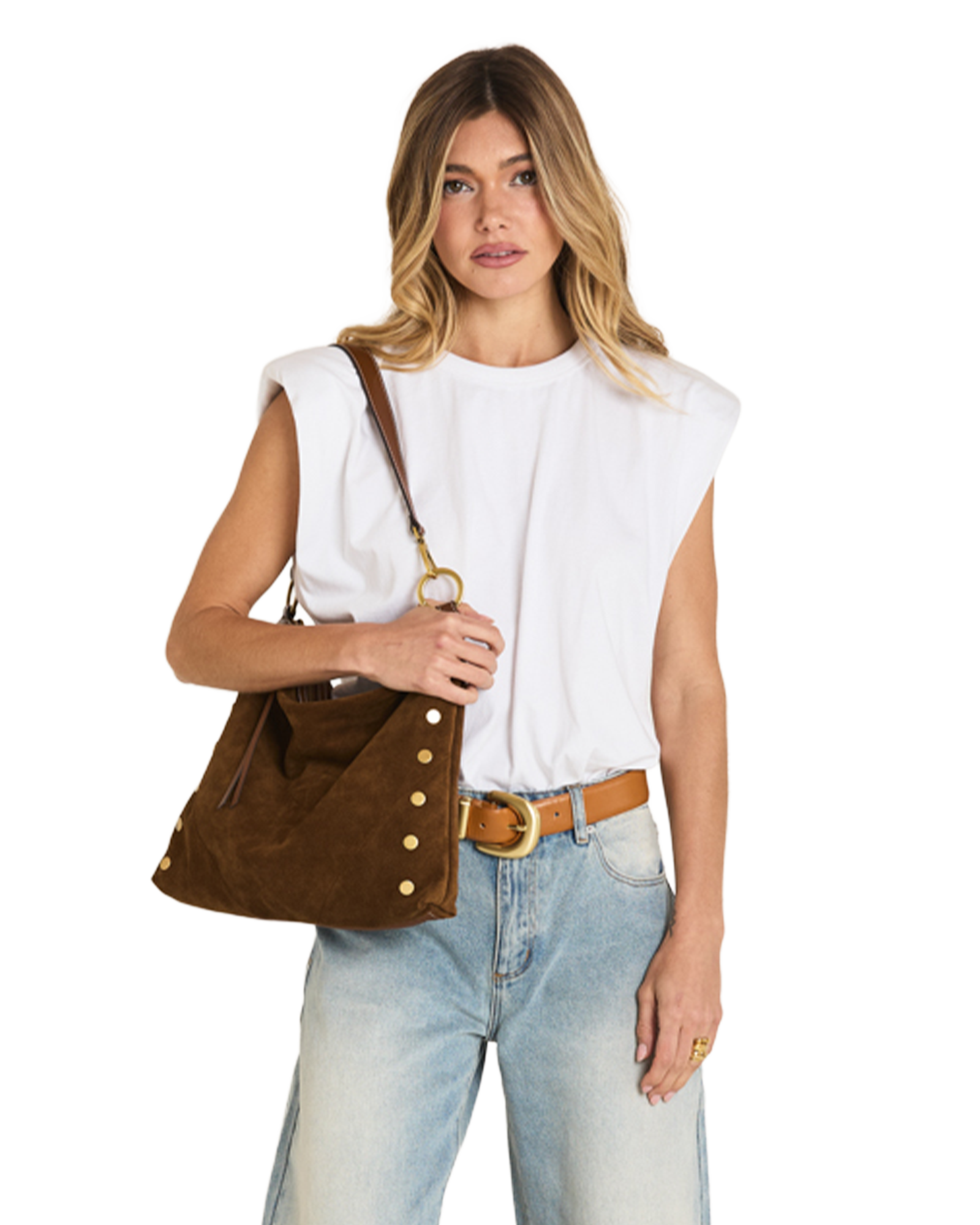 A woman modeling a brown suede shoulder bag with gold-tone studs and a leather strap, styled with a white sleeveless top, light blue jeans, and a tan belt against a plain white background.