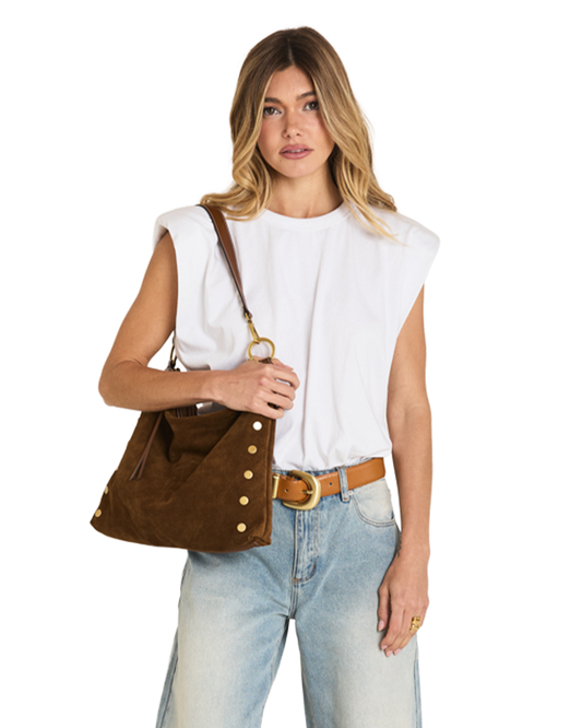 A woman modeling a brown suede shoulder bag with gold-tone studs and a leather strap, styled with a white sleeveless top, light blue jeans, and a tan belt against a plain white background.