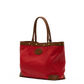 A red tote bag with brown leather handles, trim, and corner protectors, featuring brass-tone hardware and a circular brown logo patch on the front, set against a plain white background.